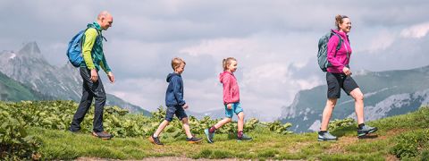 Eine vierköpfige Familie wandert auf einem Pfad in der Nähe eines Sees mit Spiegelungen im Wasser. Die Gruppe besteht aus zwei Erwachsenen und zwei Kindern, alle tragen Rucksäcke. Sie sind von üppigen grünen Hügeln umgeben und im Hintergrund ist ein bewölkter Himmel.