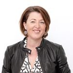 Eine Frau mit kurzen braunen Haaren lächelt in die Kamera. Sie trägt eine schwarze Lederjacke über einer gemusterten Bluse und silberne Ohrringe. Der Hintergrund ist schlicht weiß.