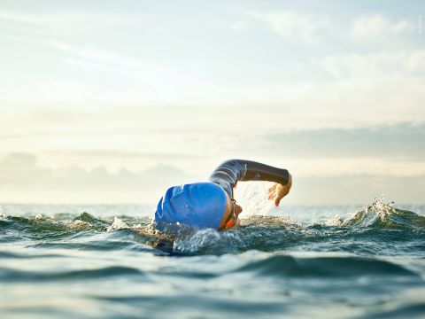 Ein Schwimmer mit blauer Badekappe und Schwimmbrille schwimmt im offenen Wasser Kraulstil. Die sonnenbeschienenen Wellen plätschern um sie herum und der Horizont ist unter einem sanften, bewölkten Himmel sichtbar.