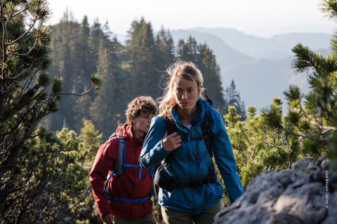 Zwei Personen wandern einen felsigen Pfad hinauf, der von üppigen grünen Bäumen umgeben ist. Die Person vorn trägt eine blaue Jacke, während die Person dahinter eine rote Jacke trägt. Im Hintergrund sind Bergkämme unter einem klaren Himmel zu sehen.