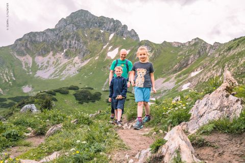 Ein Mann und zwei Kinder wandern auf einem felsigen Pfad in den Bergen. Die Kinder stehen im Vordergrund, lächeln und sind leger gekleidet. Der Mann folgt ihnen. Im Hintergrund sind grüne Hügel und ein bewölkter Himmel zu sehen.