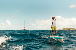 Eine Person in weißem Hemd und gelben Shorts paddelt auf dem Meer. Der Himmel ist teilweise bewölkt und in der Ferne links ist ein Segelboot zu sehen. Um die Person herum brechen sanft die Wellen.