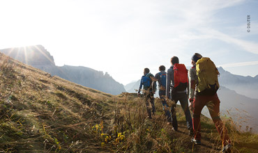 Eine Gruppe Wanderer mit Rucksäcken geht einen grasbewachsenen Hügel hinauf, im Hintergrund sieht man eine Bergkette und eine helle Sonne. Der Himmel ist klar und blau, was auf einen sonnigen Tag schließen lässt.