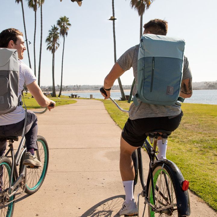 Zwei Personen fahren mit dem Fahrrad auf einem gepflasterten Weg entlang eines Parks am Wasser. Beide tragen Rucksäcke und legere Kleidung. Auf beiden Seiten des Weges sind Palmen und Gras, im Hintergrund ist das Wasser unter einem blauen Himmel zu sehen.