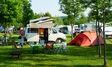 Ein Campingplatz mit einem geparkten Wohnmobil, einem orangefarbenen Zelt und mehreren Klappstühlen um einen Tisch. Fahrräder stehen in der Nähe und im Hintergrund sind zwischen grünen Bäumen und Gras weitere Wohnmobile und Fahrzeuge zu sehen.