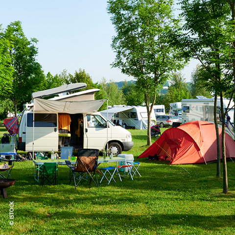 Ein heller, sonniger Tag auf einem Campingplatz mit einem weißen Wohnmobil, einem roten Zelt und mehreren Tischen und Campingstühlen. Bäume und Gras umgeben das Gebiet, und im Hintergrund sind andere Wohnmobile und Fahrzeuge zu sehen.