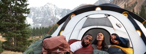 Drei Personen in Schlafsäcken stehen in einem Zelt mit geöffnetem Zelteingang und schauen nach draußen. Im Hintergrund sind sie von Bergen und Bäumen umgeben, was an eine Campingszene in einer malerischen Umgebung im Freien erinnert.