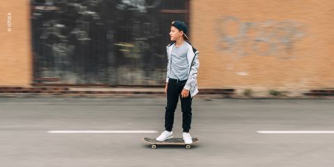 Ein kleiner Junge mit einer Kappe fährt mit Skateboard eine Straße entlang. Er trägt einen Kapuzenpullover, ein T-Shirt und Jeans. Im Hintergrund ist eine verschwommene Hauswand mit Graffiti zu sehen.
