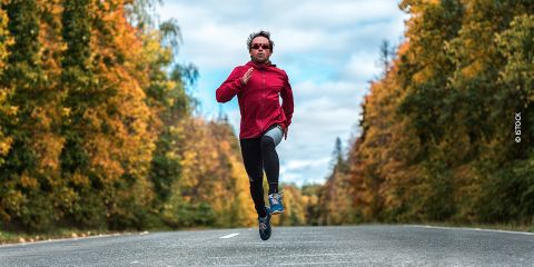 Eine Person in einer roten Jacke und schwarzen Leggings läuft auf einer leeren Straße, umgeben von Bäumen mit Herbstlaub. Sie trägt eine Sonnenbrille und der Himmel ist teilweise bewölkt.