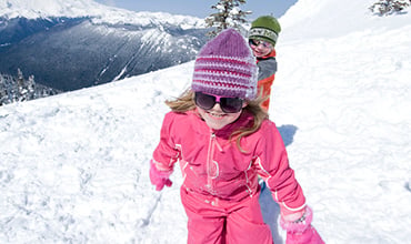 Zwei Kinder spielen an einem sonnigen Tag im Schnee und tragen bunte Winterkleidung. Das Mädchen vorne trägt einen rosa Schneeanzug mit einer lila Mütze und der Junge dahinter ein orangefarbenes Outfit mit einer grünen Mütze. Im Hintergrund sind schneebedeckte Berge zu sehen.