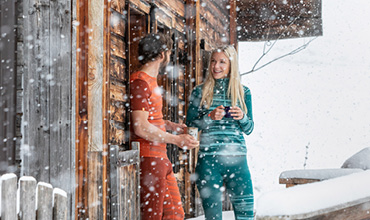 Ein Mann und eine Frau in heller Thermokleidung stehen auf der Veranda einer Holzhütte, unterhalten sich und halten Tassen in den Händen. Um sie herum fällt heftiger Schnee, der Dach und Boden mit einer dicken weißen Schicht bedeckt.