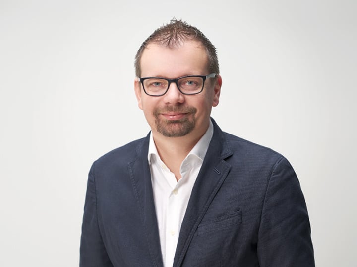 Ein Mann mit kurzen Haaren und Brille trägt einen marineblauen Blazer über einem weißen Hemd. Er lächelt und steht vor einem schlichten hellen Hintergrund.