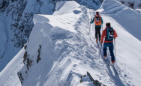 Zwei Menschen mit Rucksäcken und Winterkleidung wandern einen schmalen, schneebedeckten Bergkamm entlang. Sie stützen sich mit Stöcken ab, umgeben von steilen, schneebedeckten Gipfeln unter klarem Himmel.