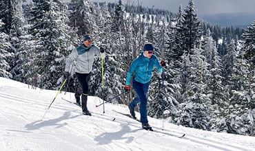 Zwei Personen laufen auf einer verschneiten Loipe Ski, umgeben von schneebedeckten Bäumen. Beide tragen Winterkleidung, darunter Jacken, Mützen und Sonnenbrillen. Der Himmel ist bedeckt und die Landschaft ist ein Winterwunderland.
