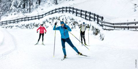 Drei Personen laufen auf einer verschneiten Loipe Ski. Der Skifahrer vorn trägt ein blaues Outfit, zwei weitere folgen ihm, einer in Rosa, der andere in Grau. Schneebedeckte Bäume und ein Holzzaun säumen den Weg.