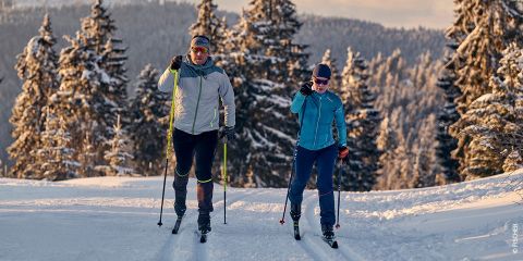 Zwei Personen laufen auf einer schneebedeckten Loipe Ski, umgeben von schneebedeckten immergrünen Bäumen. Beide tragen Wintersportkleidung und die Szene spielt vor dem Hintergrund einer sonnenbeschienenen Winterlandschaft.