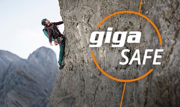 Ein Kletterer mit Helm und Ausrüstung erklimmt eine steile Felswand. Im Vordergrund steht in fetten weißen und orangefarbenen Buchstaben der Text „giga SAFE“. Im Hintergrund sind Berggipfel zu sehen.