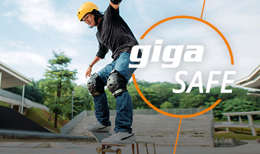 Eine Person mit gelbem Helm und Knieschützern fährt im Freien mit dem Skateboard eine Rampe hinunter. Die Worte „giga SAFE“ sind deutlich daneben zu sehen. Im Hintergrund sind Bäume und ein Gebäude zu sehen.