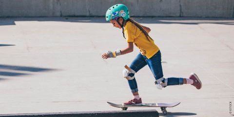 Ein junges Mädchen mit blaugrünem Helm, gelbem Hemd, Jeans und Schutzkleidung fährt in einem Skatepark Skateboard. Sie ist in Bewegung, stößt sich mit einem Fuß ab und balanciert mit dem anderen auf dem Brett. Der Hintergrund ist eine Betonfläche.