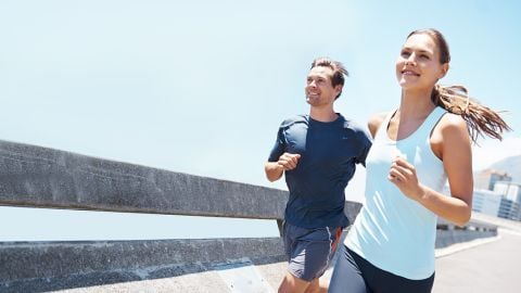 Ein Mann und eine Frau joggen an einem sonnigen Tag. Sie lächeln und tragen Sportkleidung. Sie laufen über eine Brücke mit einer Stadtlandschaft im Hintergrund und einem klaren blauen Himmel über ihnen.