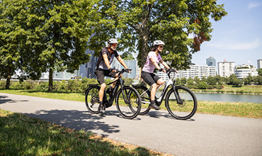 Zwei Personen fahren auf einem gepflasterten Weg in einem Park mit großen Bäumen am Himmel Rad. Im Hintergrund sind ein Gewässer und die Skyline einer Stadt zu sehen. Beide Radfahrer tragen Helme und legere Radsportkleidung und genießen einen sonnigen Tag.