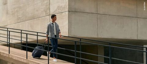 Person, die auf einem Betonweg geht und einen Seesack mit Rollen hinter sich herzieht. Sie trägt eine graue Jacke, blaue Jeans und weiße Schuhe. Im Hintergrund ist ein modernes Betongebäude mit klaren Linien und einem einfachen, minimalistischen Design zu sehen.