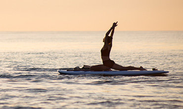 Eine Person in einer Yoga-Pose auf einem Paddleboard auf dem Meer bei Sonnenuntergang. Die Silhouette wird von den warmen Farben des Abendhimmels eingerahmt, umgeben von ruhigem Wasser.