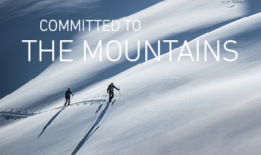 Zwei Menschen wandern unter klarem Himmel auf einem schneebedeckten Berghang. Der Schnee ist makellos, man sieht deutliche Spuren hinter ihnen. Oben steht der Text „Committed to the Mountains“.