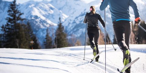 Zwei Personen laufen auf Skiern auf einer verschneiten Loipe, umgeben von immergrünen Bäumen und Bergen unter einem klaren blauen Himmel. Sie tragen Winterkleidung und sind von hinten zu sehen, wie sie durch die malerische Landschaft laufen.