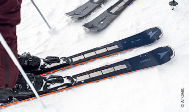 Nahaufnahme eines Paars Skier im Schnee, in dessen Nähe eine Person steht. Die Skier sind dunkel mit hellen Kanten und haben ein sichtbares Markenlogo. Andere Skier sind teilweise im Hintergrund sichtbar.