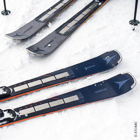 Nahaufnahme mehrerer Paar schwarzer Skier mit orangefarbenen Akzenten, die auf dem Schnee liegen. Auf den Skiern ist der Markenname „Atomic“ zu sehen.