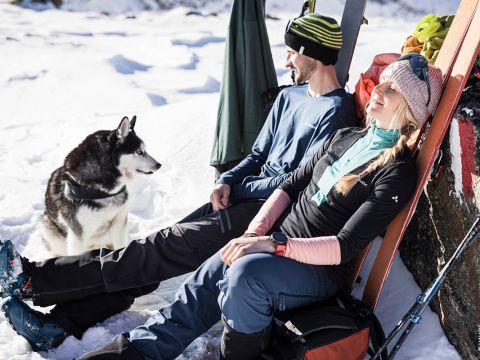 Ein Mann und eine Frau in Winterkleidung entspannen vor einer verschneiten Kulisse. Skier stehen bereit. Die Frau lächelt mit geschlossenen Augen, während ein Husky vor ihnen sitzt. Helles Sonnenlicht beleuchtet die ruhige, verschneite Szenerie.