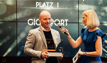 Ein Mann mit einer Auszeichnung auf der Bühne wird von einer Frau mit Mikrofon interviewt. Im Hintergrund ist vor glitzerndem Hintergrund der Schriftzug „PLATZ 1 GIGASPORT“ zu sehen.