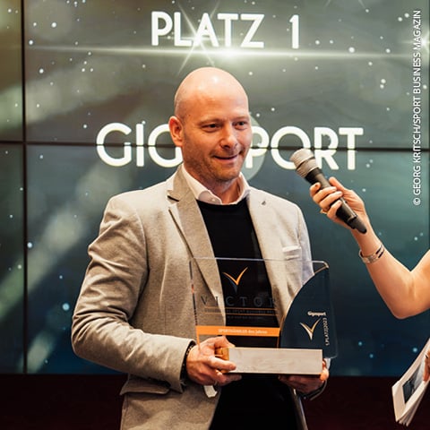 Eine Person im Anzug hält auf der Bühne eine Auszeichnung in die Höhe, neben ihr ein Mikrofon. Hinter ihr leuchtet ein Bildschirm mit den Aufschriften „PLATZ 1“ und „GIGASPORT“. Der dunkle Hintergrund mit hellen Punkten sorgt für eine festliche Atmosphäre.