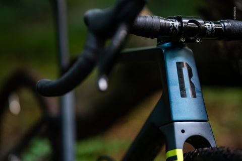 Nahaufnahme der Vorderseite eines Fahrrads mit metallisch blauem Rahmen, schwarzem Lenker mit strukturiertem Griff und markantem schwarzen Logo. Der Hintergrund ist verschwommenes Grün und lässt an eine Außenkulisse denken.