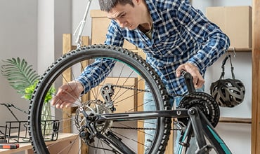 Eine Person im karierten Hemd beugt sich über ein Fahrrad und stellt das Hinterrad ein oder untersucht es. Die Szene scheint in einem Innenbereich zu sein. Im Hintergrund sind verschiedene Werkzeuge, eine Topfpflanze und Helme zu sehen.