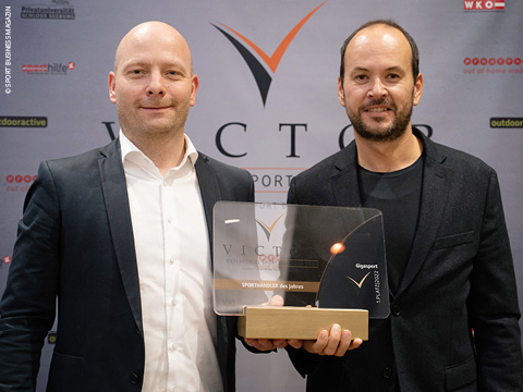 Zwei Personen in formeller Kleidung stehen nebeneinander vor einem Hintergrund mit mehreren Logos. Eine Person hält eine Plakette oder Trophäe mit der Aufschrift „Victor Sportmanager des Jahres“. Beide lächeln in die Kamera.