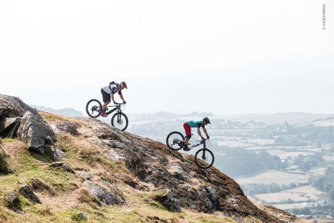 Zwei Radfahrer fahren einen felsigen Hügel in einer zerklüfteten Landschaft hinunter. Sie tragen Helme und Radsportbekleidung und fahren hintereinander her. Im Hintergrund ist eine neblige Aussicht auf Hügel und ein Tal zu sehen.