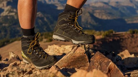 Eine Person in schwarzen Wanderschuhen mit gelben Schnürsenkeln steht auf felsigem Gelände. Im Hintergrund sind Berge unter klarem Himmel unscharf zu sehen.