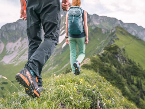 Zwei Wanderer überqueren einen grasbewachsenen Bergkamm. Im Vordergrund sind die Trekkingschuhe eines Wanderers in Nahaufnahme zu sehen. Beide tragen Rucksäcke und sind von üppig grünen Hügeln und einem bewölkten Himmel umgeben.