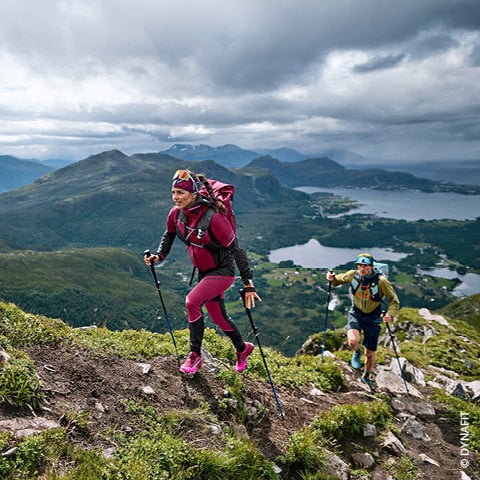 Zwei Wanderer mit Rucksäcken und Trekkingstöcken steigen einen grasbewachsenen Bergpfad hinauf. Der Wanderer im Vordergrund trägt Rosa und Lila, der dahinter Grün und Blau. Unter einem dramatischen Wolkenhimmel bietet sich ein malerischer Blick auf Täler, Seen und ferne Berge.