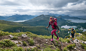 Zwei Personen wandern mit Trekkingstöcken einen üppigen Bergpfad hinauf. Die Landschaft ist geprägt von grünen Hügeln, fernen Bergen und einem bewölkten Himmel, der einen malerischen Blick auf Täler und Seen bietet. Der führende Wanderer ist rosa gekleidet, gefolgt von einem anderen in Gelb.