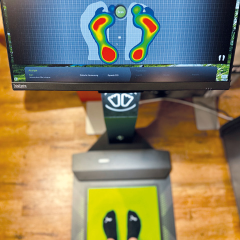 Ein Fußscanner zeigt eine Heatmap-Analyse des Fußdrucks einer Person auf dem Bildschirm. Das Scan-Gerät steht auf einem Holzboden. Darauf steht eine Person, die schwarze Socken mit kleinen weißen Symbolen trägt.