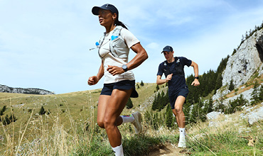 Zwei Personen laufen Trailrunning auf einem schmalen, grasbewachsenen Pfad, umgeben von Hügeln und Bäumen. Sie tragen Sportkleidung und Mützen, ihre Trinkrucksäcke sind gut sichtbar. Der Himmel ist klar, was auf einen sonnigen Tag schließen lässt.