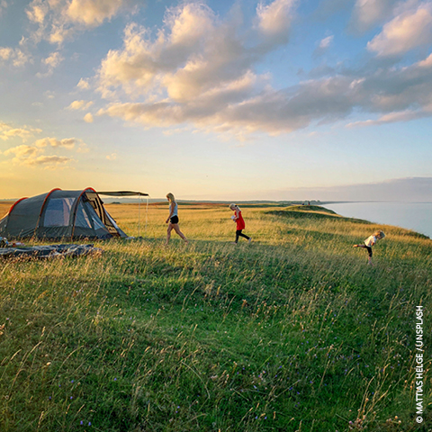 Eine Gruppe von Menschen, zwei Erwachsene und ein Kind, geht auf ein Zelt auf einem grasbewachsenen Hügel mit Blick aufs Meer zu. Der Himmel ist teilweise bewölkt, die Sonne schimmert warm. Die Landschaft ist offen und malerisch.