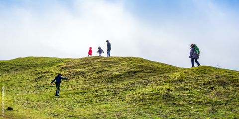 Eine Gruppe von Menschen, darunter ein Kind in einer roten Jacke, geht unter strahlend blauem Himmel auf einem grasbewachsenen Hügel spazieren. Sie sind entlang des Hangs verstreut, einige klettern hinauf, andere gehen oben entlang. Die Landschaft ist lebendig und offen.