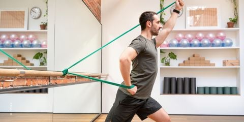 Ein Mann im Fitnessstudio benutzt ein Widerstandsband, das an einer Ballettstange befestigt ist. Er befindet sich in Ausfallschrittposition und zieht das Band mit angewinkelten Armen. Im Raum stehen Regale mit Fitnessgeräten und Pflanzen.
