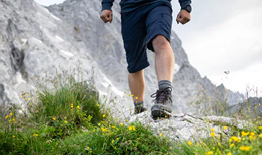 Eine Person in Shorts und Wanderschuhen steht auf einem felsigen Untergrund, umgeben von grünem Gras und kleinen gelben Blumen, im Hintergrund steile, graue Berge. Der Himmel ist bedeckt.