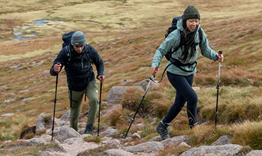 Zwei Personen wandern einen felsigen Pfad bergauf. Sie benutzen Trekkingstöcke und tragen Winterkleidung, darunter Mützen und Jacken. Im Hintergrund ist Grasland mit Felsbrocken zu sehen. Das Wetter scheint klar und frisch.