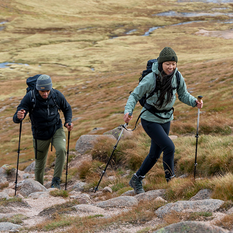 Zwei Personen wandern einen felsigen Pfad bergauf. Sie tragen Jacken, Mützen und Rucksäcke und benutzen Trekkingstöcke. Die Landschaft ist grasbewachsen mit vereinzelten Felsen und Erdflecken. Im Hintergrund ist eine weite, offene Fläche unter bewölktem Himmel zu sehen.